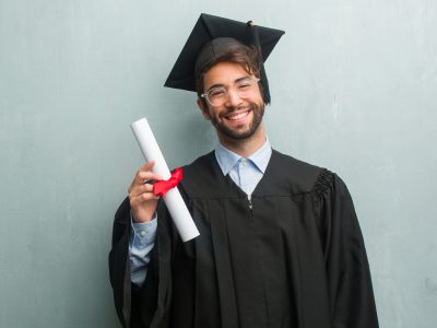 Young graduated man against a grunge wall with a copy space cheerful and with a big smile, confident, friendly and sincere, expressing positivity and success